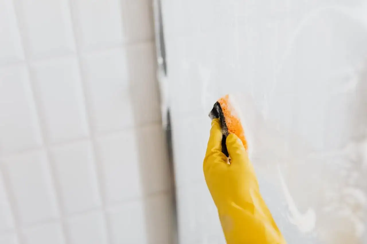 Mano con guante amarillo limpiando la mampara con esponja. Consejos para limpiar las gomas de la mampara y dejarla reluciente.