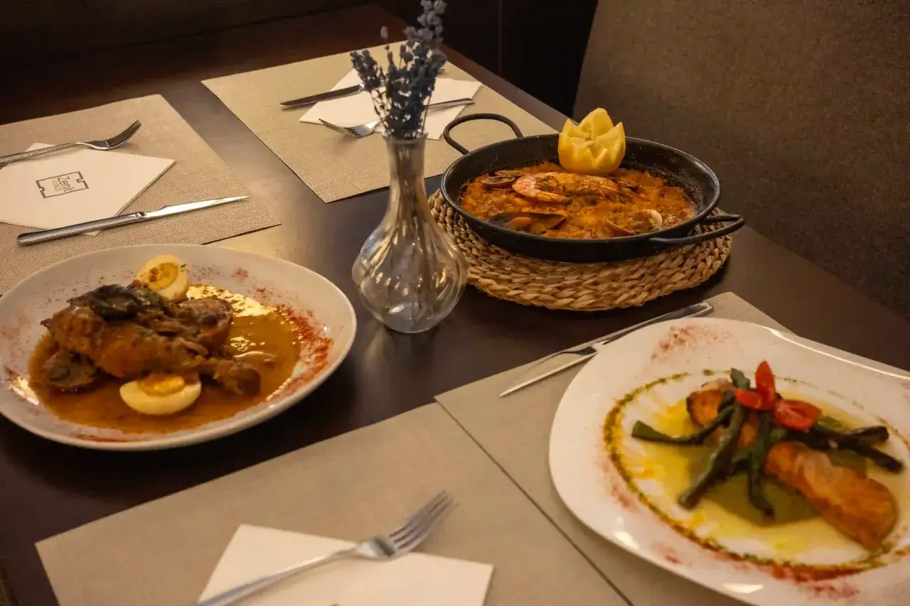 Una mesa con paella, salm&oacute;n y un plato de carne con huevo duro. La paella tiene gambas y mejillones, como si fueran roquetas de mar verde esmeralda.