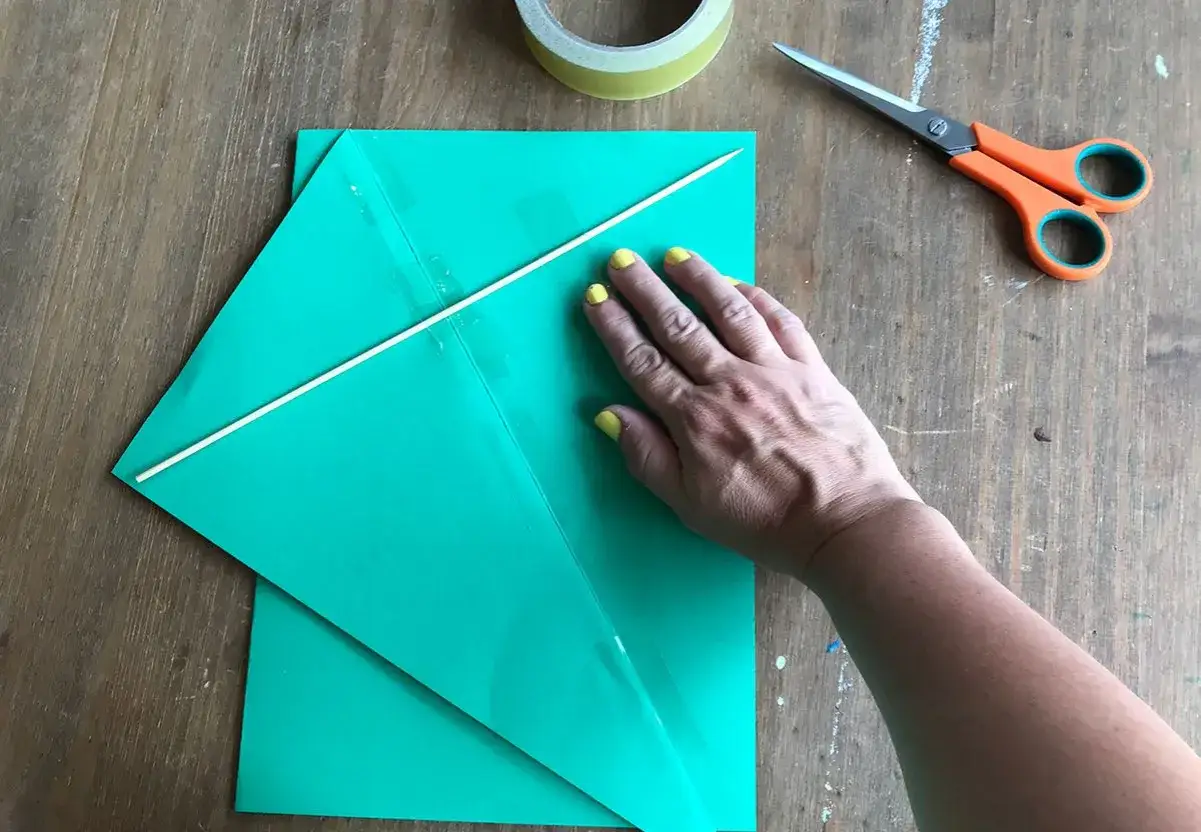 Ręka z ż&oacute;łtym lakierem przytrzymuje zielony papier, tworząc latawiec diy. Obok nożyczki i taśma.