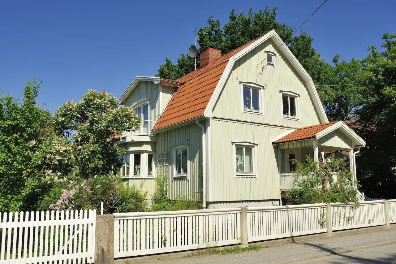 Ljusgrönt trähus med tegeltak och vit staket. En vacker trädgård med blommande buskar ramar in huset, perfekt för att byta fasad och ge huset nytt liv.