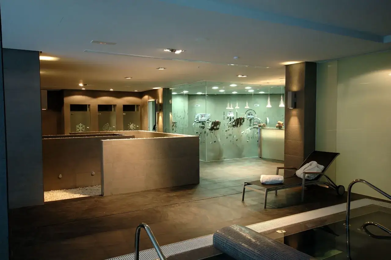 Un spa primus moderno con piscina, tumbona con toallas y zona de gimnasio con máquinas de ejercicio tras un cristal.