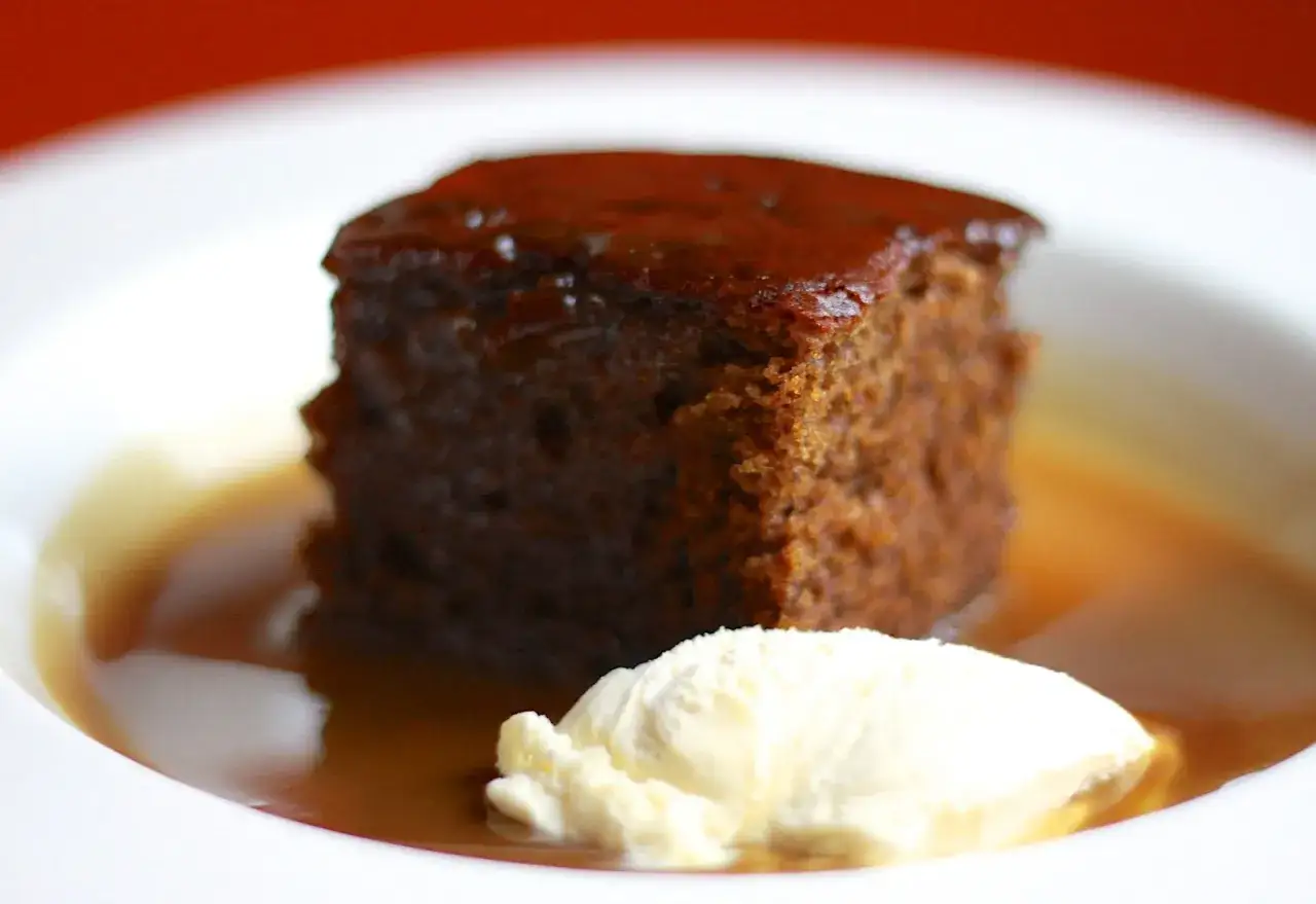 Wilgotny, ciemny kawałek ciasta sticky toffee polany sosem, z gałką lodów waniliowych obok.