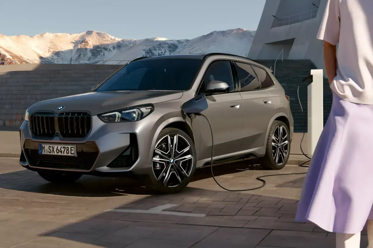 Szare BMW hybryda podłączone do ładowarki, na tle gór i nowoczesnego budynku.