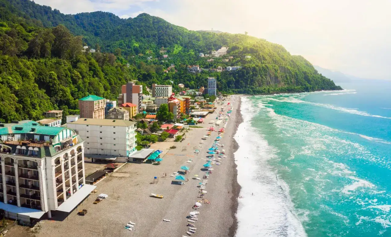 Widok na plażę i miasto Kobuleti w Gruzji. Zielone wzgórza schodzą do turkusowego morza, a nad kamienistą plażą wznoszą się kolorowe budynki.