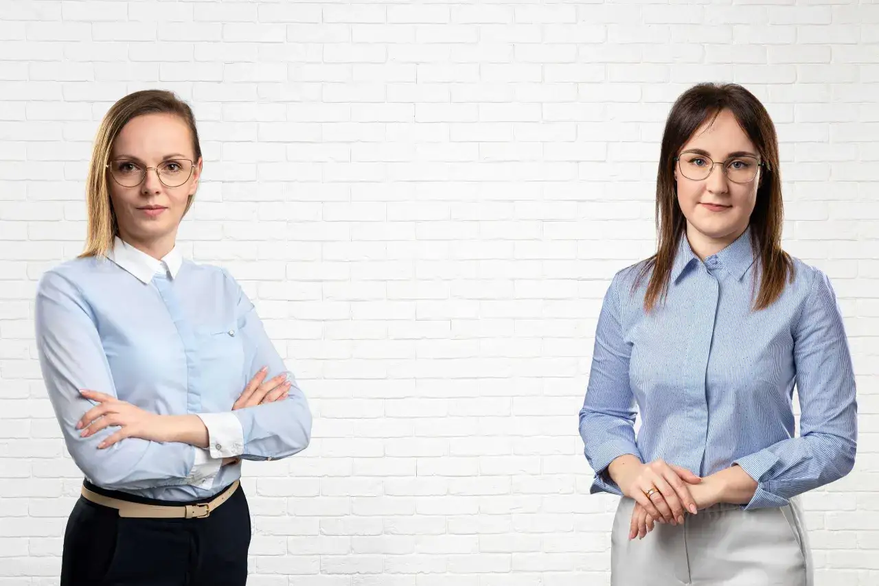 Dwie kobiety w eleganckich koszulach stoją na tle białej ceglanej ściany.