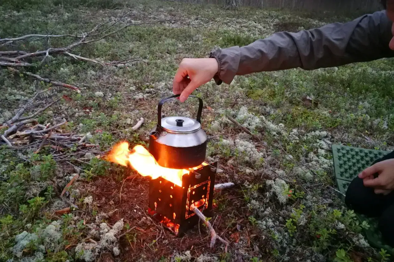 En hand lyfter en kaffepanna över en sprakande eld på en campingplats. Perfekt för friluftsmat recept.