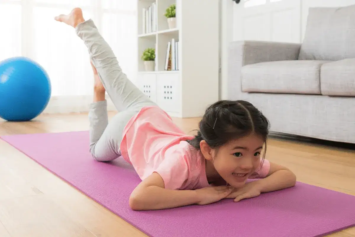 Kleines M&auml;dchen &uuml;bt Yoga im Kindergarten. Sie liegt auf einer lila Matte, ein Bein nach oben gestreckt.