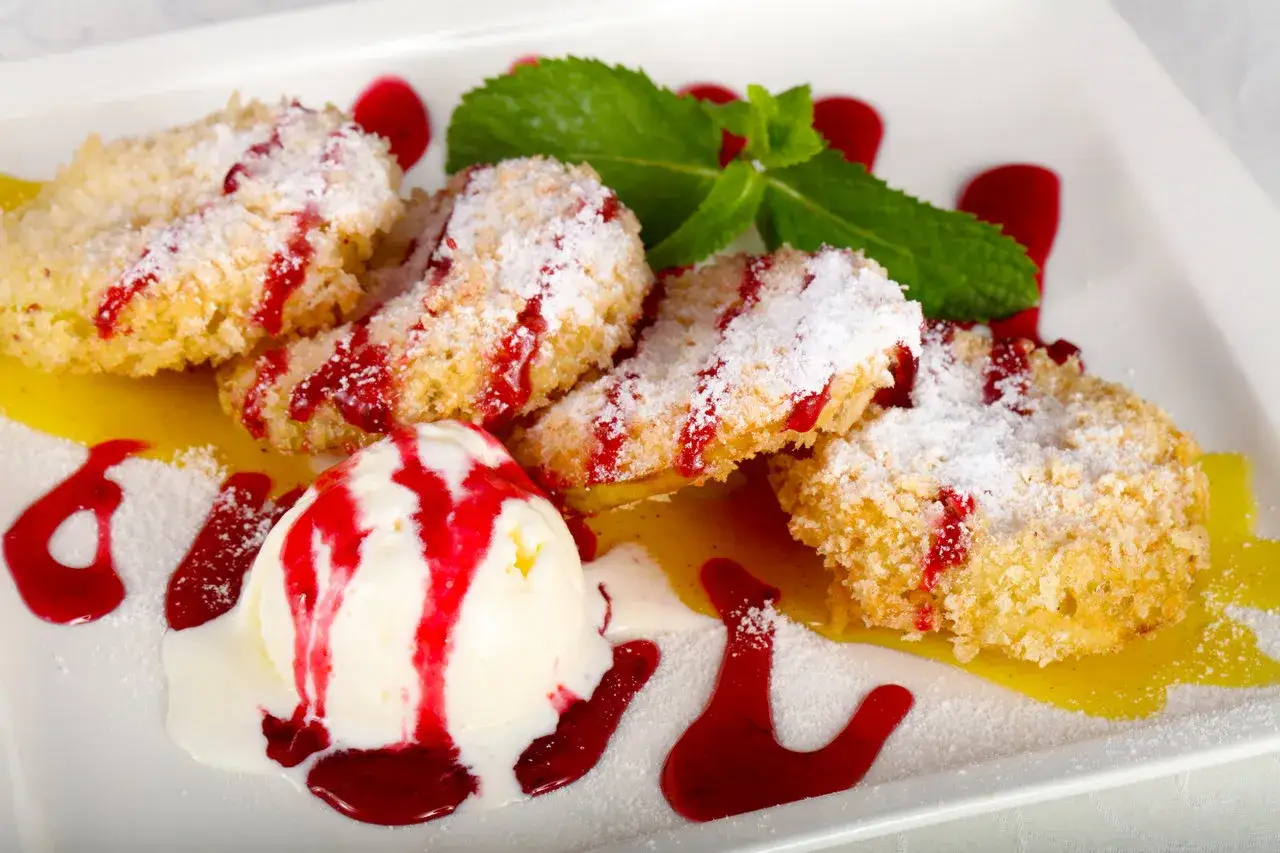 Chrupiące ciastka funnel cake posypane cukrem pudrem, polane sosem malinowym, z gałką lodów waniliowych i listkiem mięty.