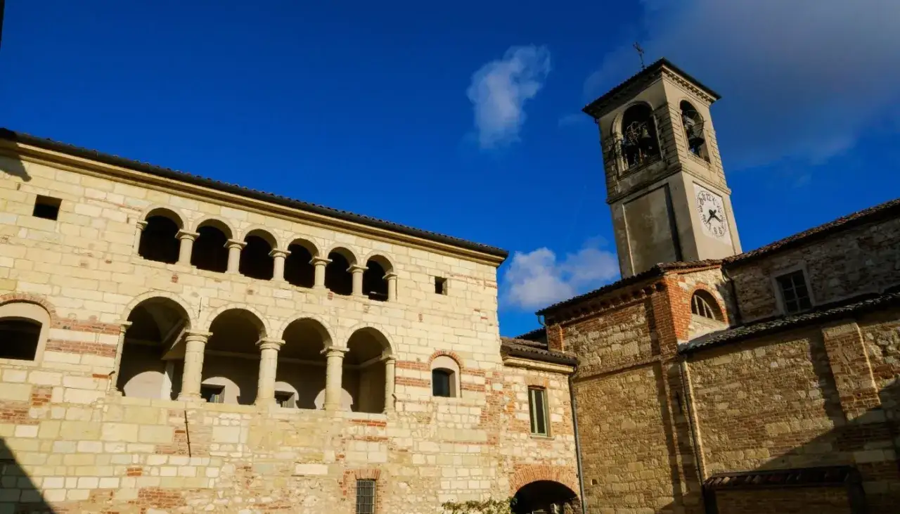 Antica **cella monte** con portici ad archi e campanile con orologio, sotto un cielo azzurro intenso.