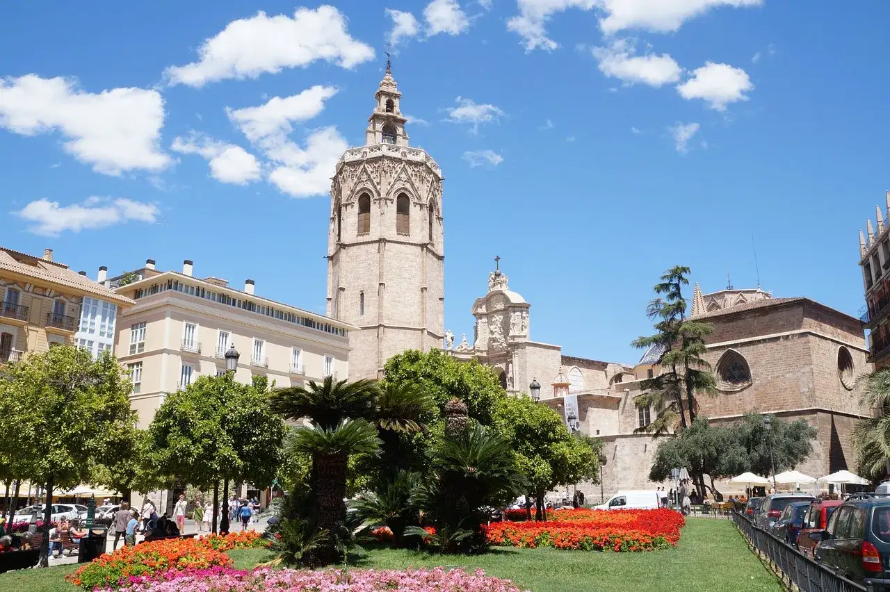 La **catedral de Valencia** se alza majestuosa bajo un cielo azul con nubes. Un jard&iacute;n florecido y &aacute;rboles verdes rodean la plaza.