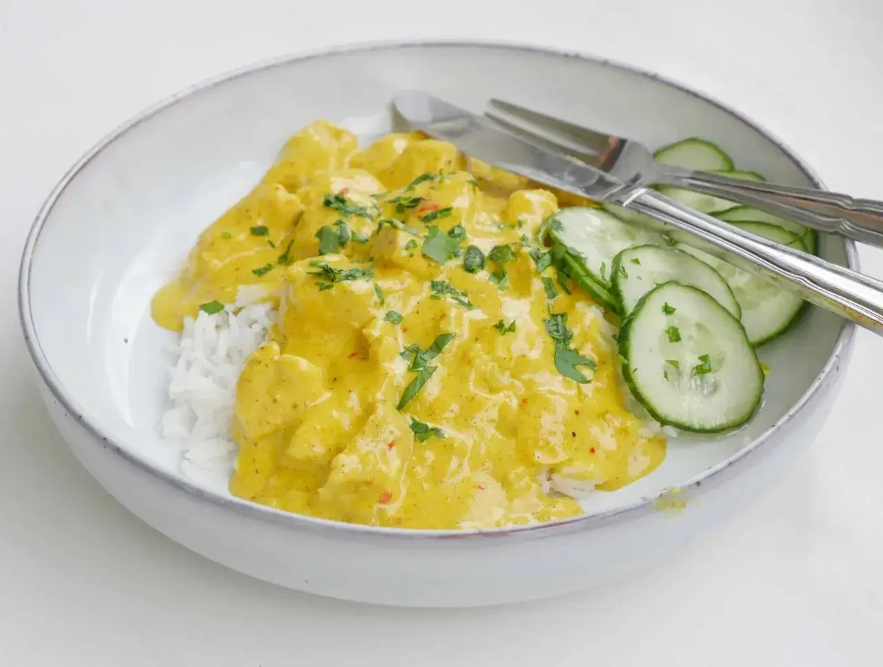 En skål med ris täckt av krämig kyckling currysås, garnerad med färsk persilja och skivad gurka. En utsökt måltid!