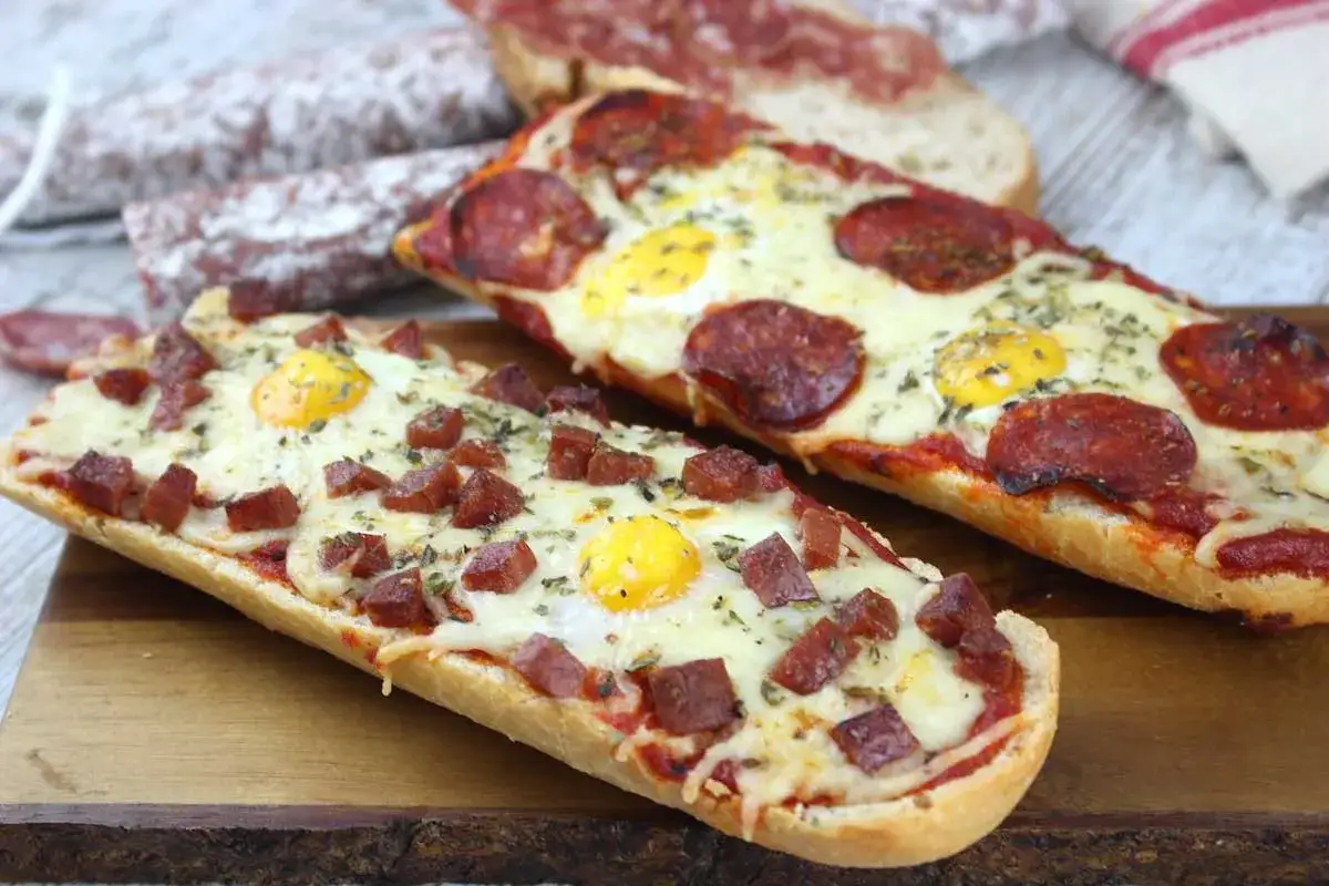 Dos pizzas caseras sobre pan alargado, una con trocitos de fuet y huevo, la otra con pepperoni y huevo.