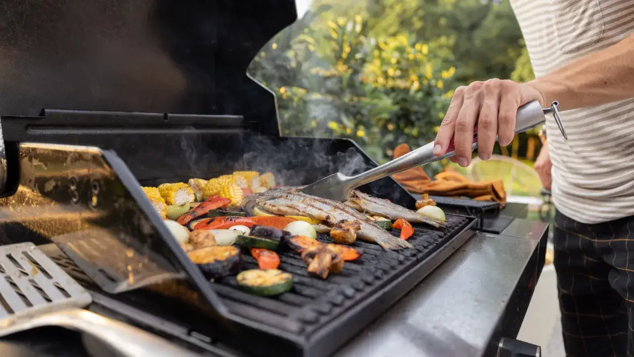 Un homme utilise une pince pour retourner du poisson et des légumes sur un barbecue à gaz.