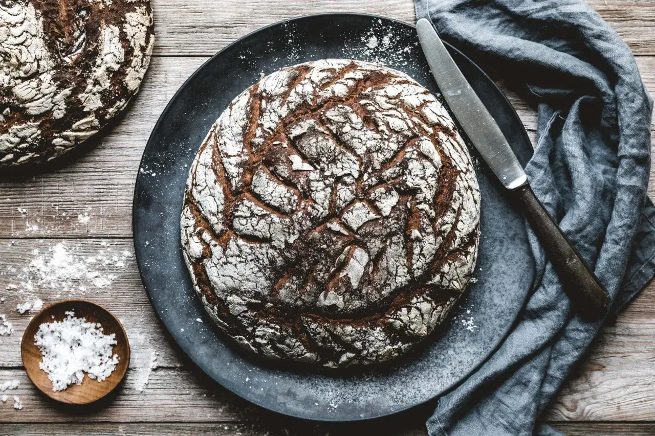 Ein rustikales Roggenbrot mit Sauerteig liegt auf einem dunklen Teller, daneben ein Messer und eine kleine Schale mit Salz.