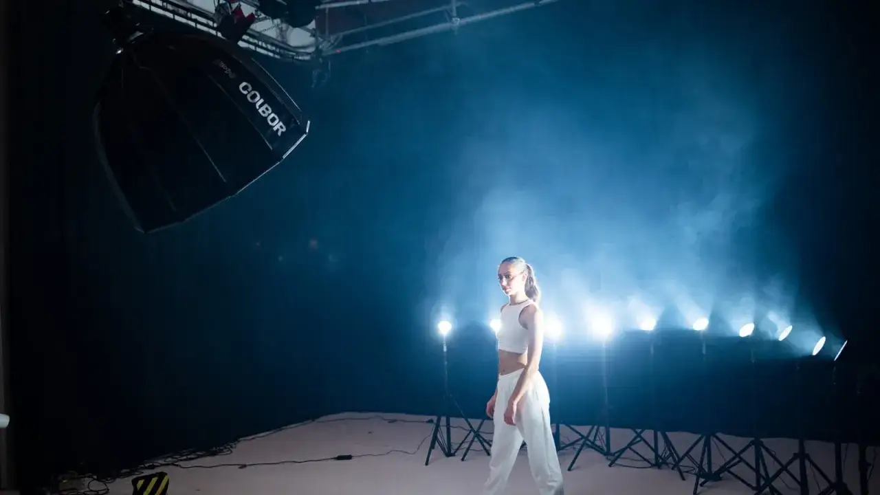 A young woman in white athletic wear poses in a smoky studio during a fashion photography session, illuminated by bright lights.