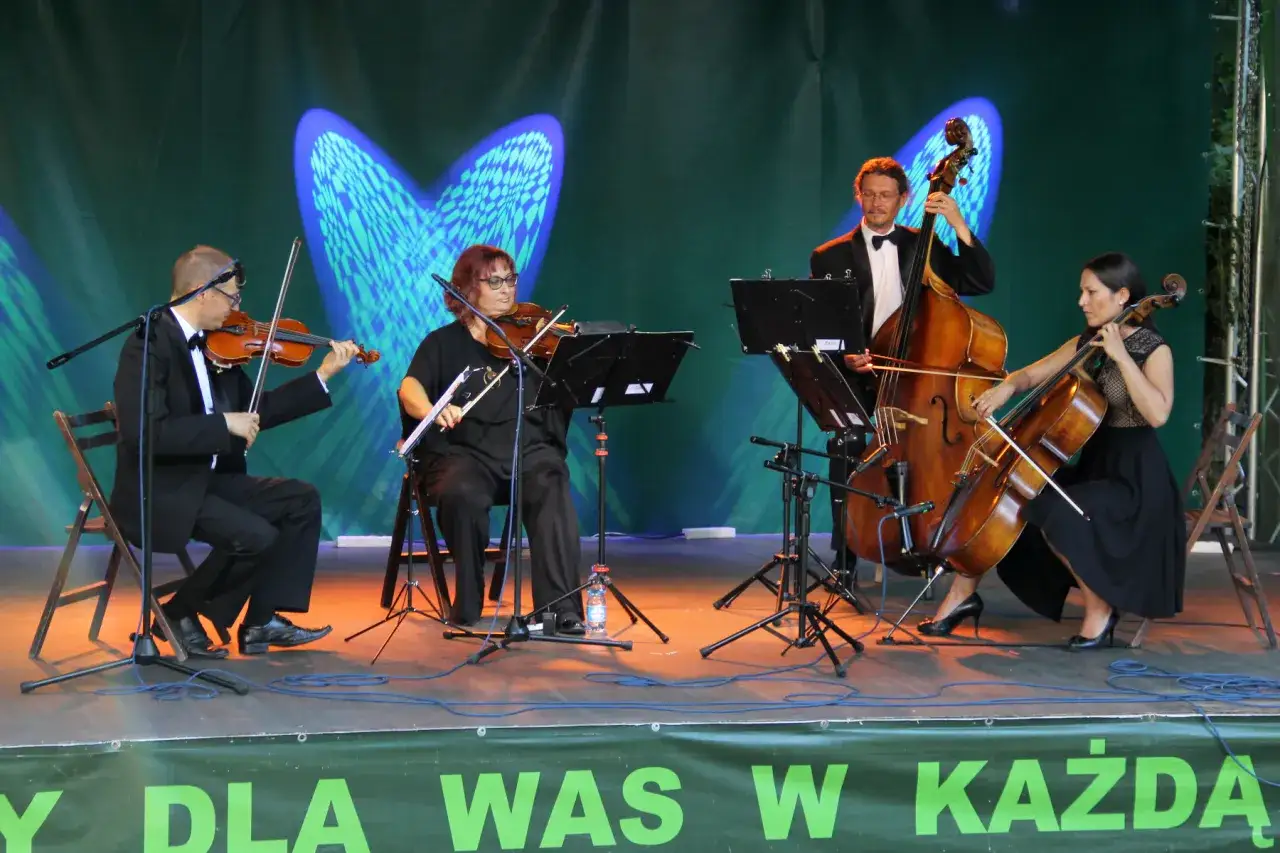 Kwartet smyczkowy gra na scenie. Muzycy w eleganckich strojach wykonują utwór na skrzypcach, altówce i kontrabasie.