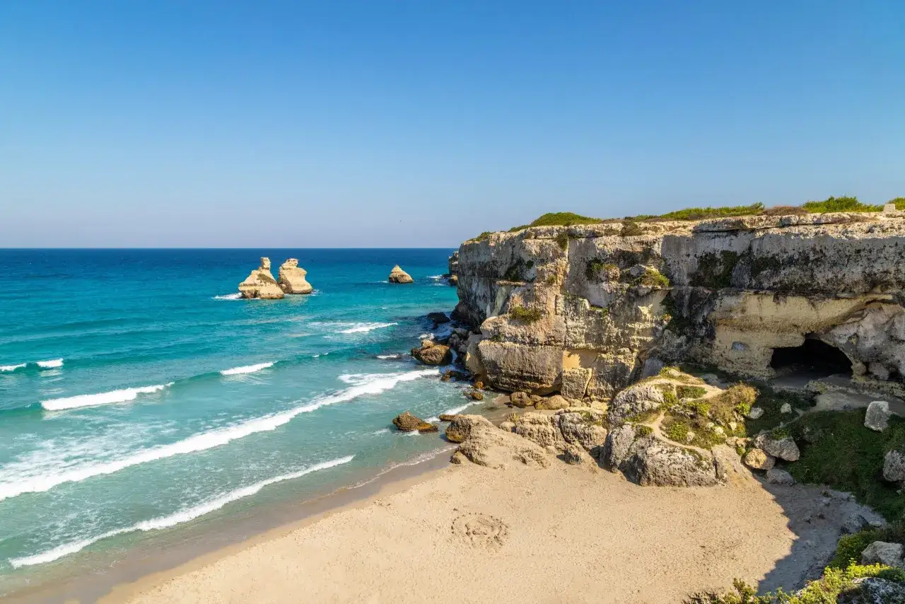 Scogliere calcaree e mare turchese in una splendida **Puglia estate**. Faraglioni emergono dall'acqua cristallina, mentre una grotta si apre nella roccia.