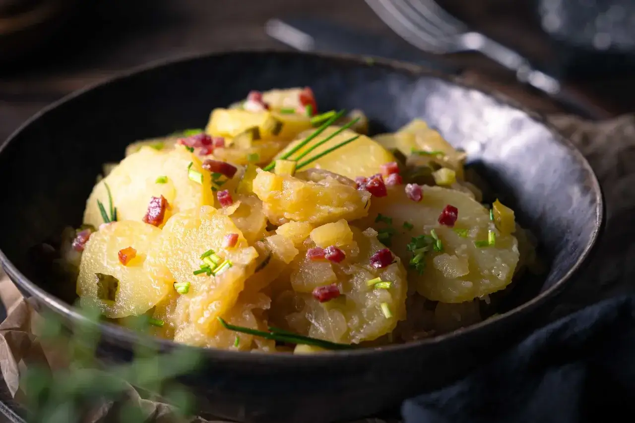Ein Teller mit warmem Kartoffelsalat, garniert mit Speckwürfeln und Schnittlauch.