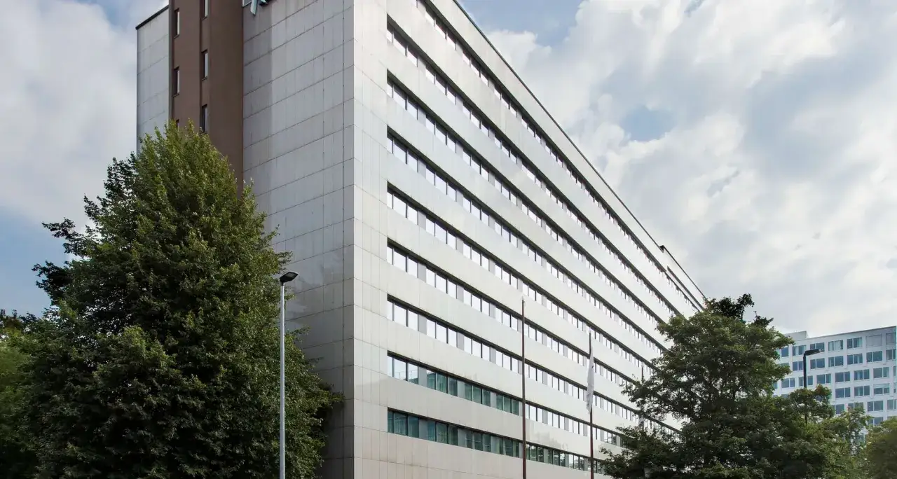 Nowoczesny budynek hotelu Radisson Blu Scandinavia Hotel w Düsseldorfie, z białymi panelami i licznymi oknami, otoczony zielenią.