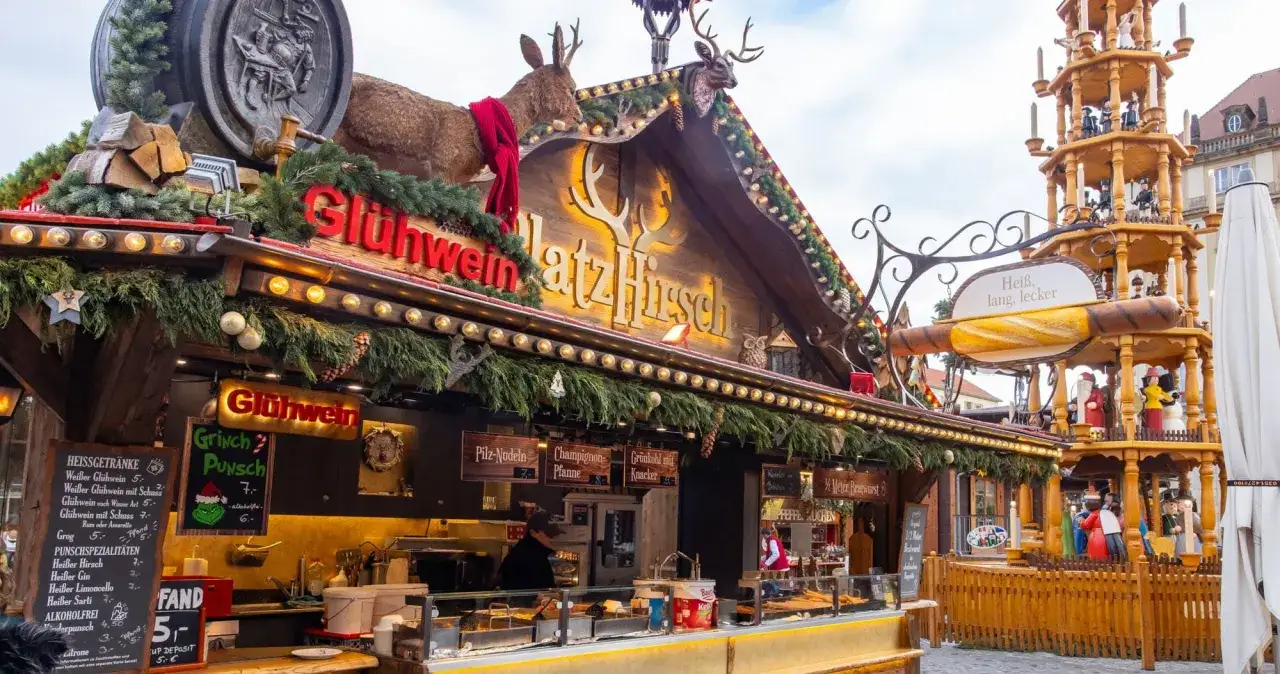 A festive Dresden Christmas market stall, adorned with fir branches and deer, offers Gl&uuml;hwein and other treats. A wooden pyramid stands nearby.