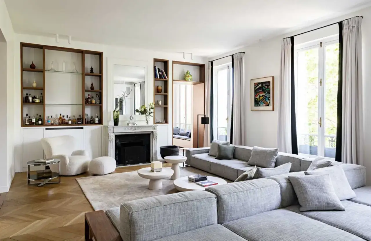 Salon lumineux avec un grand canapé gris, une cheminée, des étagères remplies de bouteilles et un fauteuil blanc. La maison de rêve parfaite pour se détendre.