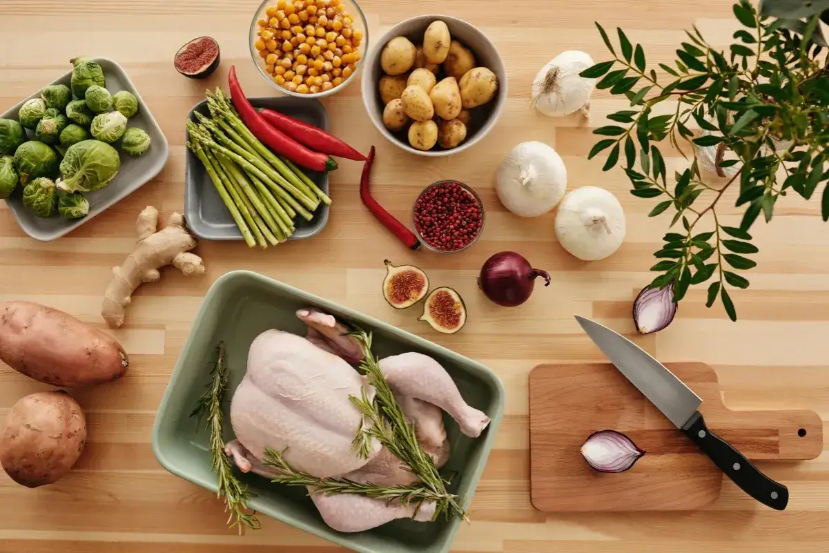 En rå kyckling ligger i en ugnsform med rosmarin. Runtom finns grönsaker som brysselkål, sparris, chili, potatis, sötpotatis, ingefära, vitlök, lök och fikon. Undrar hur smakar dålig kyckling?