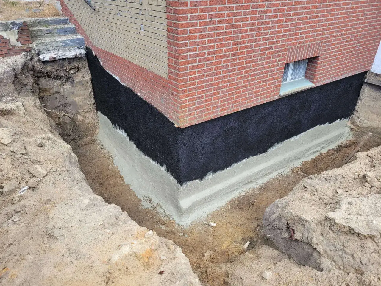 Das Fundament wird für die Kellerabdichtung vorbereitet. Schwarze Bitumenbahnen und eine graue Schutzschicht schützen vor Feuchtigkeit.