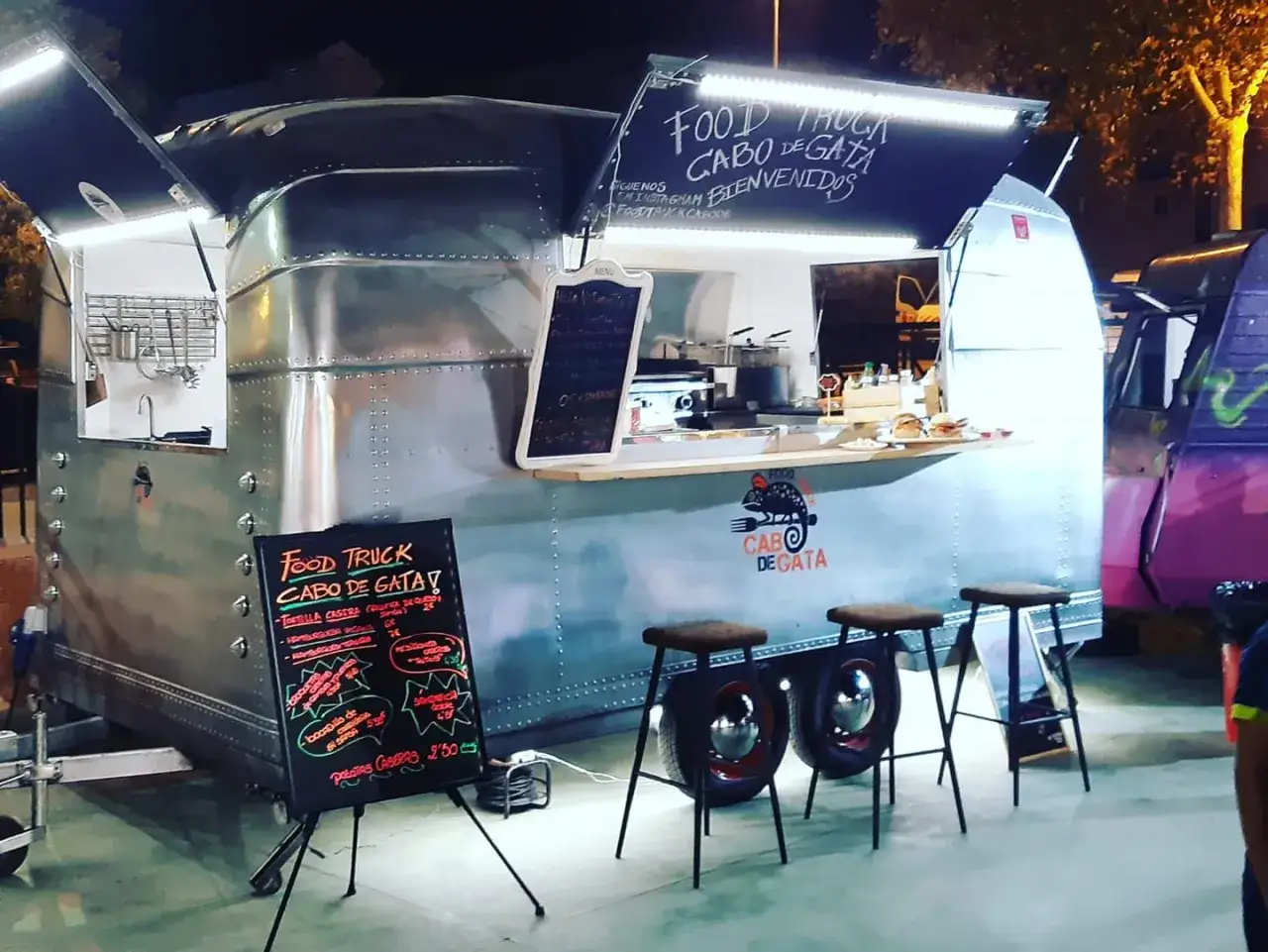 Food truck Cabo de Gata, un remolque plateado con luces brillantes, sirve hamburguesas y patatas fritas. Tres taburetes esperan a los clientes.