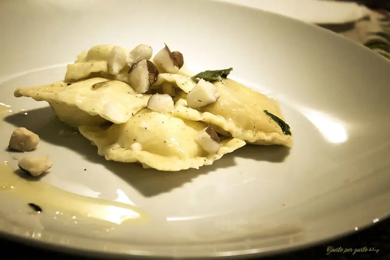 Ravioli pere e pecorino, conditi con nocciole tritate e una foglia di salvia, su un piatto bianco con un filo d'olio.