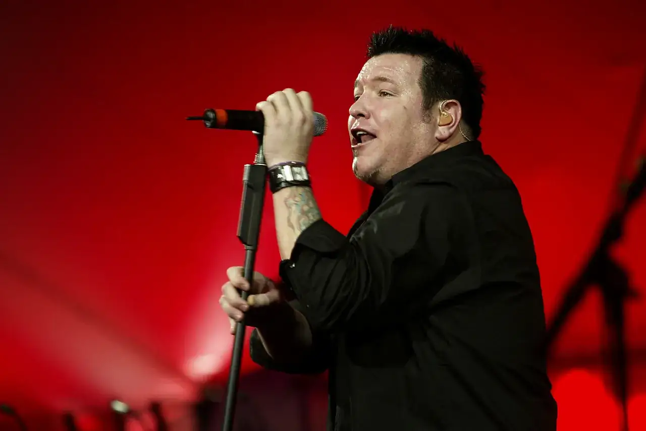 Steven Harwell, wokalista zespołu Smash Mouth, śpiewa do mikrofonu na tle czerwonej sceny.