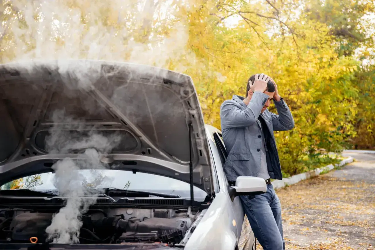 Un homme désespéré devant sa voiture en panne, le capot ouvert laissant échapper une épaisse fumée blanche. Cause surchauffe moteur essence.