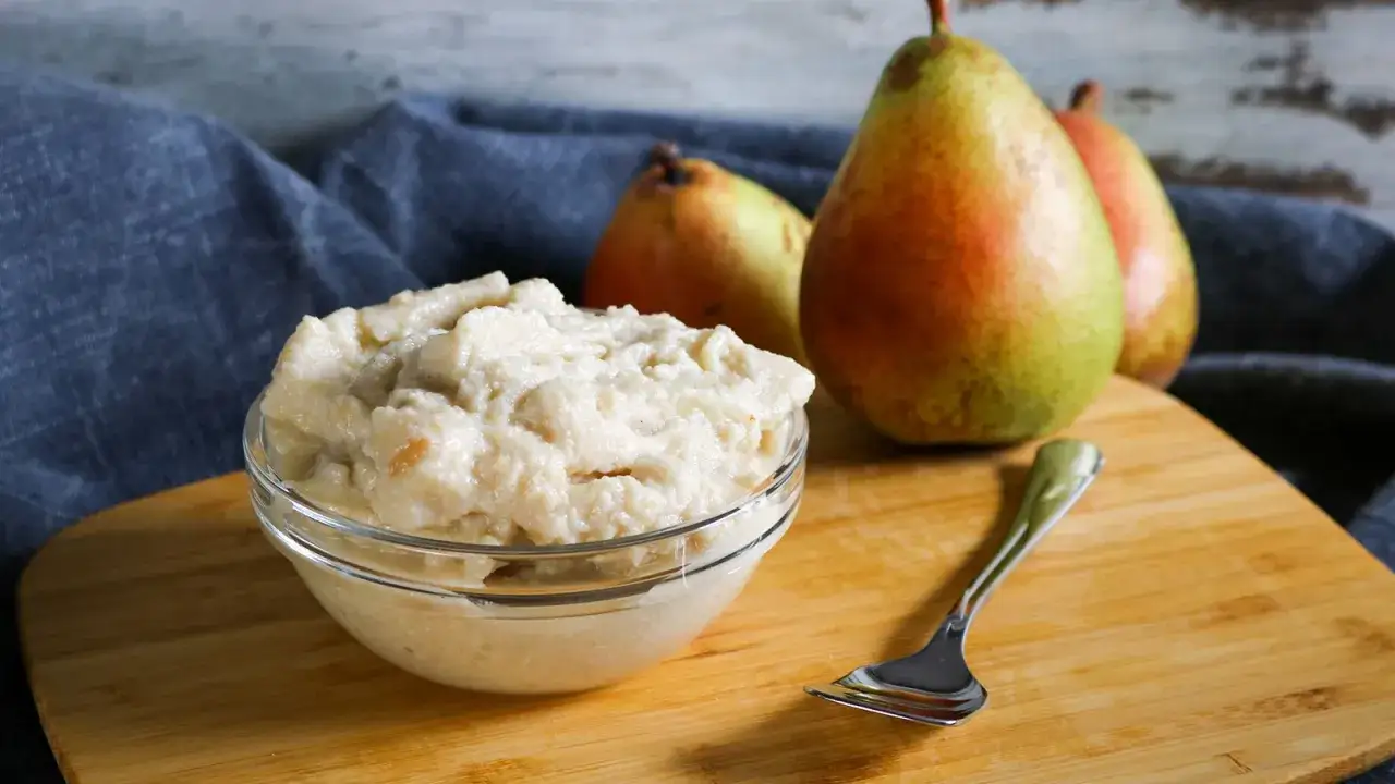 Cremiges Birnen-Eis in einer Sch&uuml;ssel, daneben reife Birnen und eine Gabel auf einem Holzbrett.