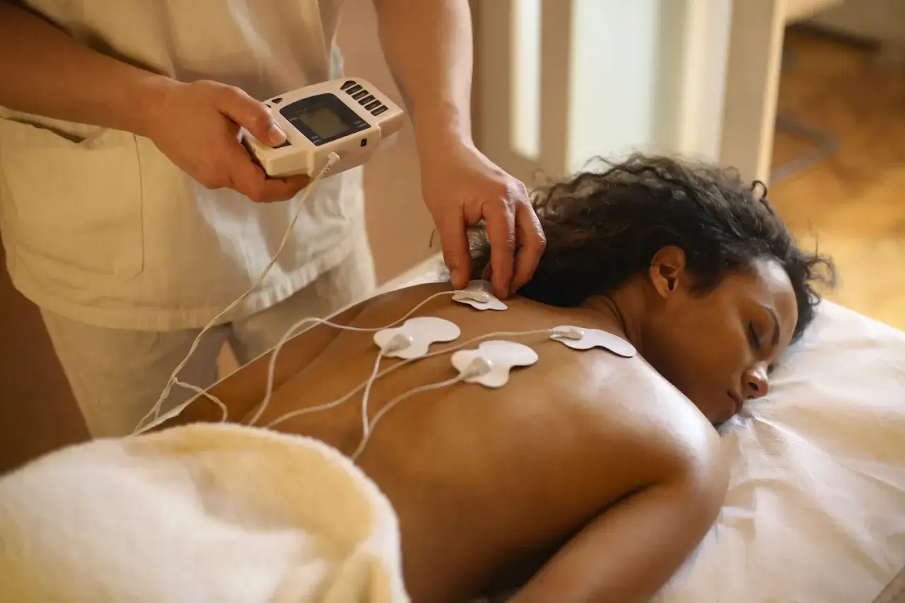 Kobieta leży na masażowym stole, a terapeuta podłącza elektrody do jej pleców. Zabieg elektrostymulacji EMS przynosi ulgę.