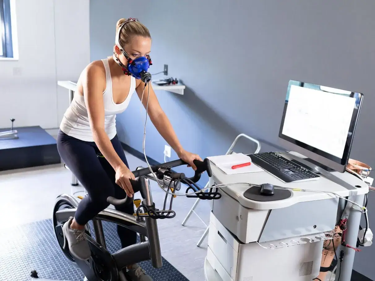 Femme en masque respiratoire effectuant un test d'effort vélo, surveillée par un ordinateur.