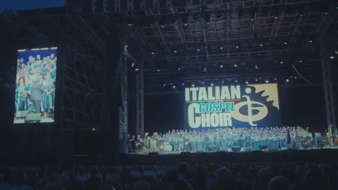 Un grande coro si esibisce sul palco, illuminato da un maxi schermo che mostra il logo 