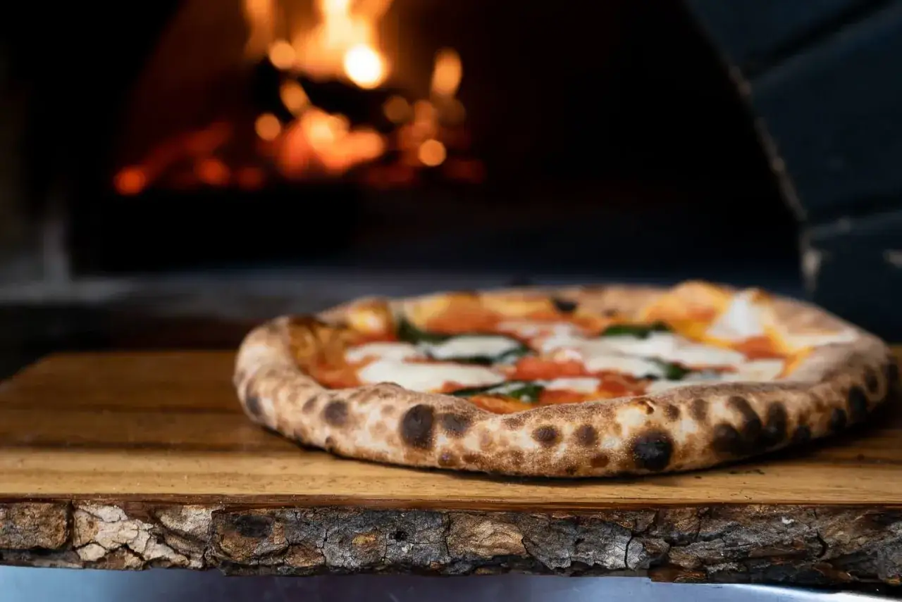Frisch gebackene Pizza Margherita auf Holzbrett vor einem Pizzaofen. K&ouml;stliche Pizza-Sorten wie diese sind ein Genuss.