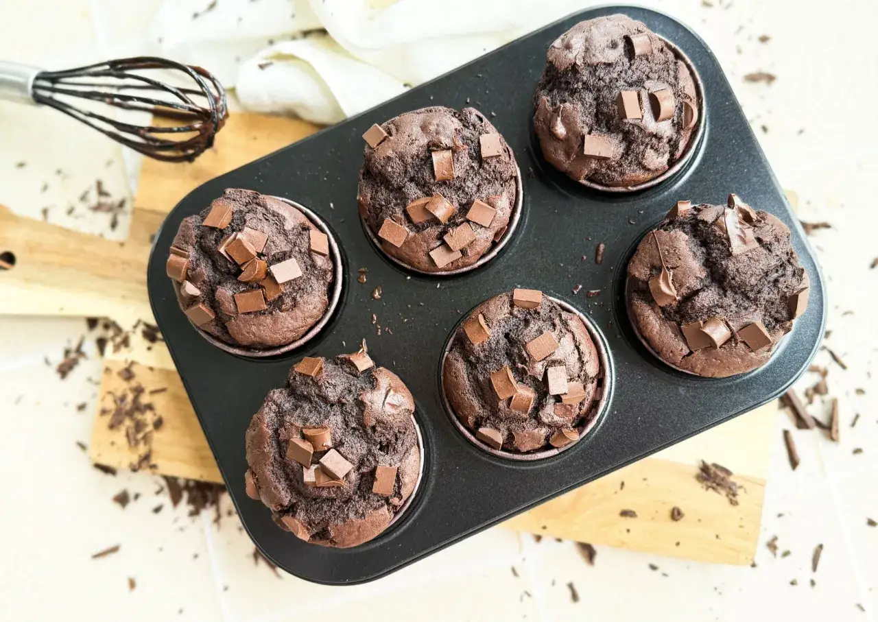 Schokoladen-Muffins ohne Ei, frisch aus dem Ofen, mit Schokostückchen garniert. Ein Schneebesen mit Schokospuren liegt daneben.