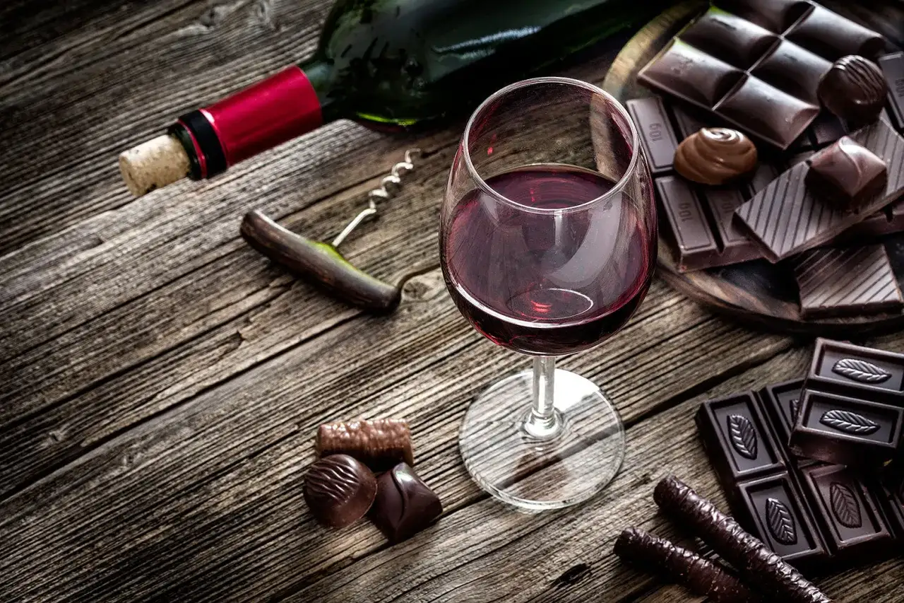 Ein Glas Rotwein und verschiedene Schokoladenst&uuml;cke auf rustikalem Holz. Ein Korkenzieher liegt neben einer Weinflasche.
