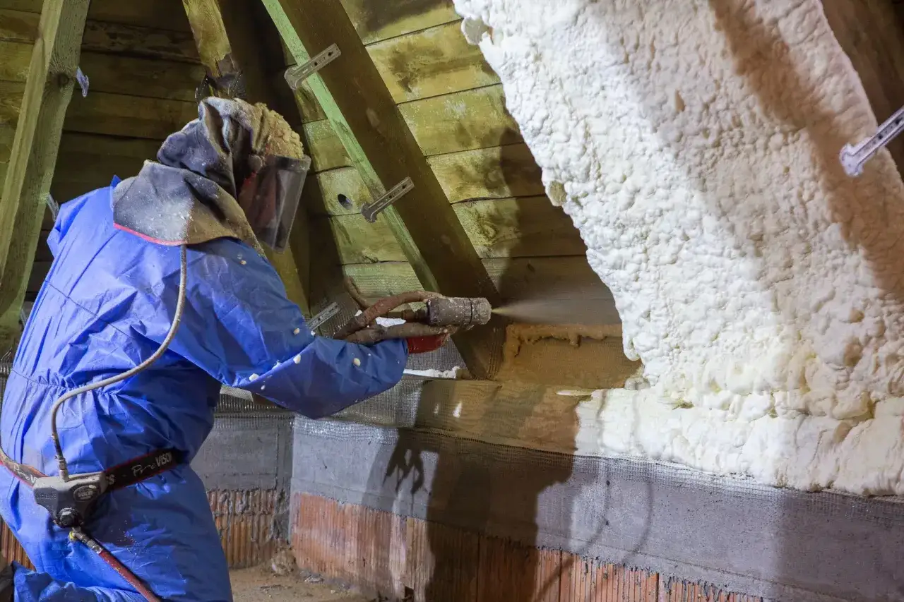 Un technicien en combinaison bleue applique du polyur&eacute;thane projet&eacute; sur les combles pour une isolation efficace.