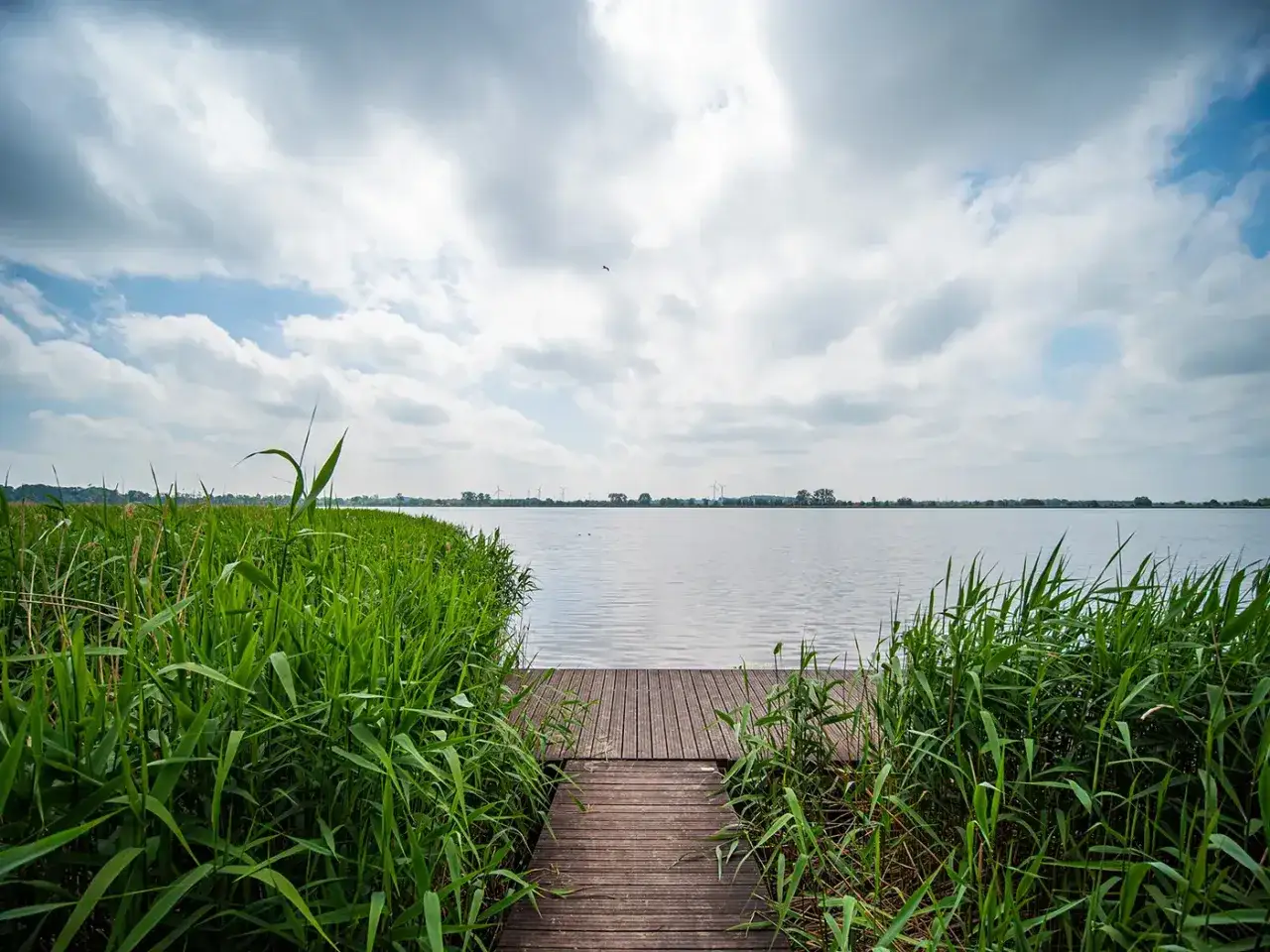 Drewniany pomost prowadzi do spokojnego jeziora, otoczonego bujną, zieloną roślinnością. W oddali widać wiatraki, a nad nimi skalno-niebieskie niebo z chmurami.