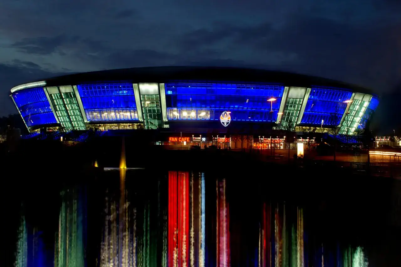 Donbass Arena nocą, oświetlona niebieskimi i zielonymi światłami, z odbiciem w wodzie.