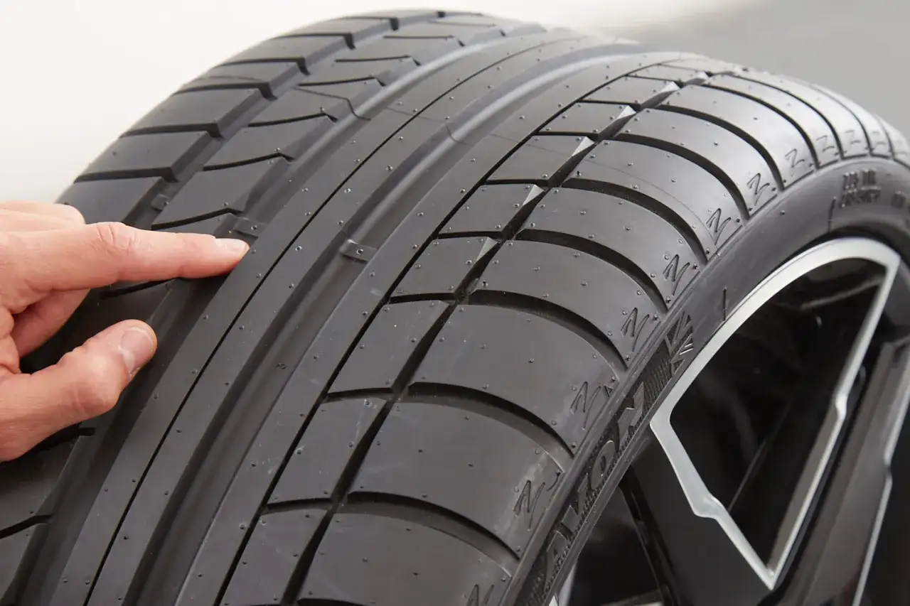 Un dedo señala la banda de rodadura de un neumático, mostrando su diseño y textura. Estos son los neumáticos, esenciales para la tracción y seguridad del vehículo.