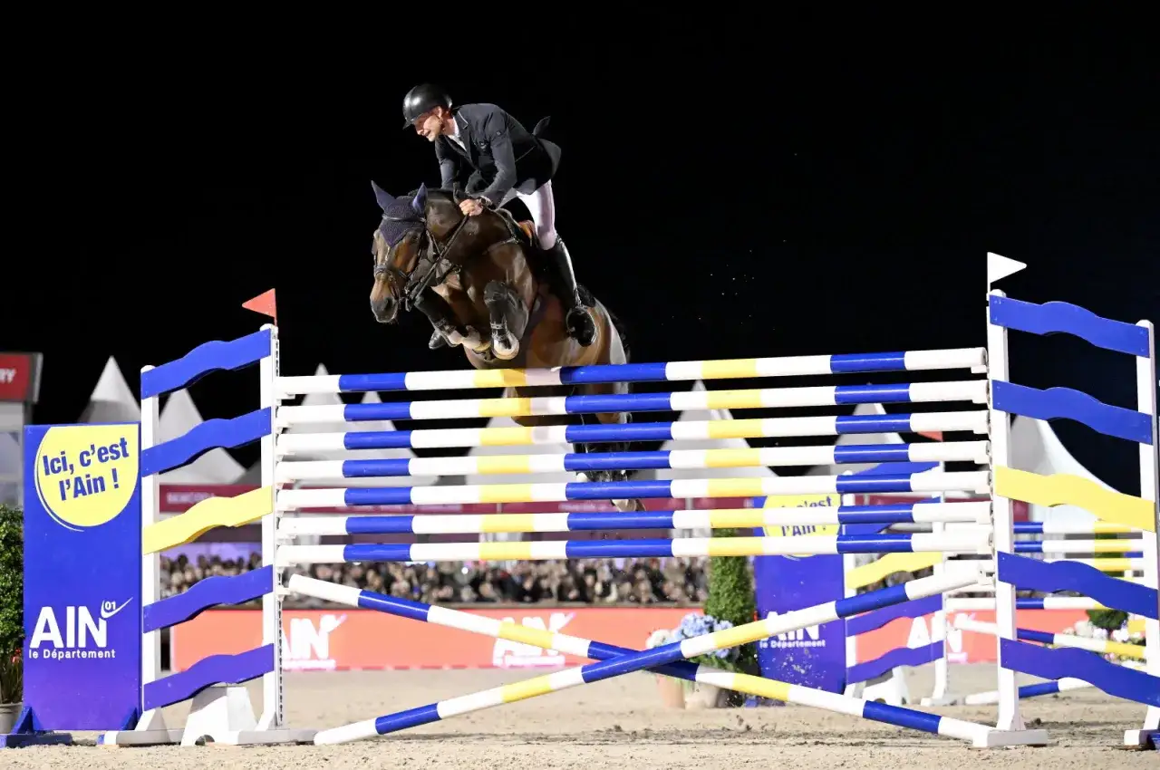 Cavalier et cheval en plein saut lors de l'épreuve des 6 barres. Le cheval est au sommet de son effort, les pattes repliées, franchissant l'obstacle.