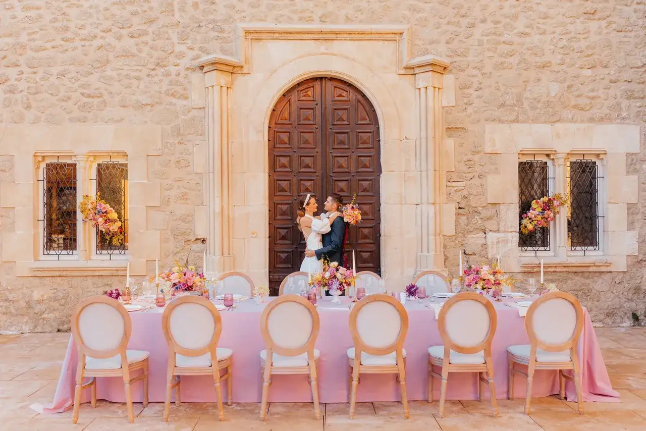 Un couple s'embrasse devant une porte massive, une table dress&eacute;e pour un festin gourmand, orn&eacute;e de fleurs roses et de bougies.