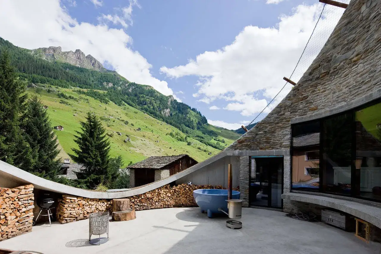 Dekorativ gestapeltes Brennholz umrahmt eine Terrasse mit Bergblick. Ein Grill und eine blaue Wanne laden zum Entspannen ein.