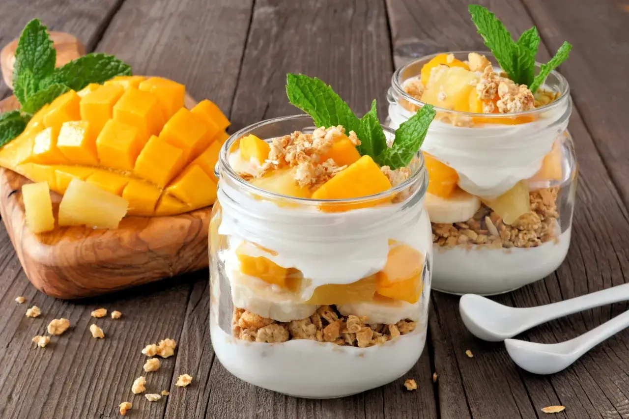 Dwa słoiki pysznych lod&oacute;w jogurtowych z mango, bananem i granolą, udekorowane miętą. Obok pokrojone mango na drewnianej desce.