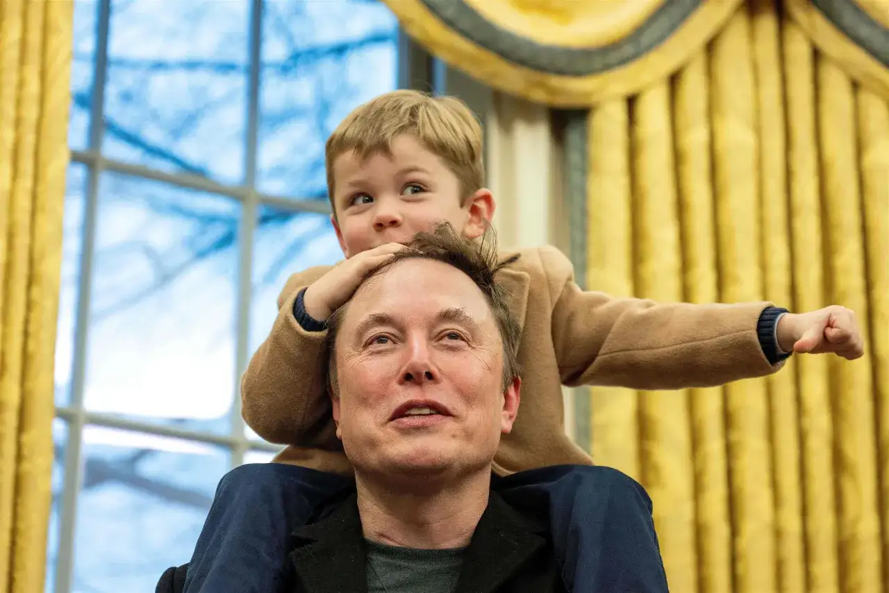 Elon Musk z synem na ramionach. Chłopiec w beżowym płaszczyku wyciąga rączkę, a Elon patrzy w górę z uśmiechem.