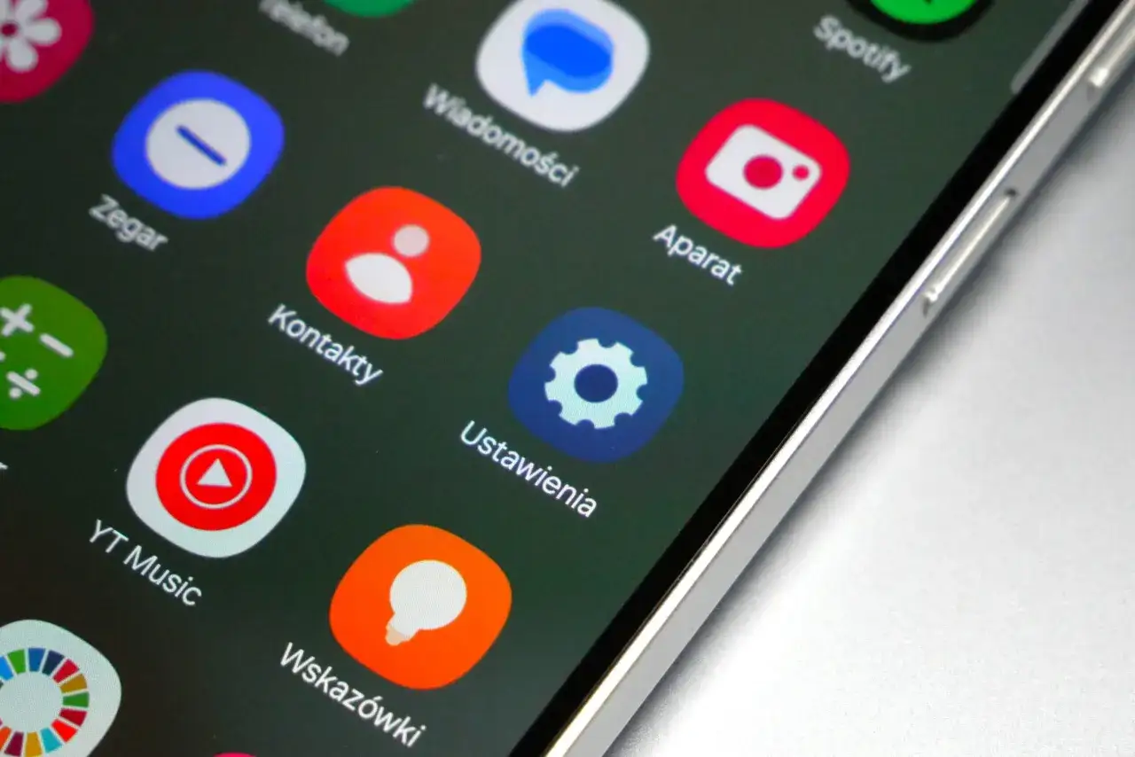 Ekran smartfona z ikonami aplikacji, w tym Zegar, Kontakty, Ustawienia, Aparat, YT Music. Widoczny fragment interfejsu One UI 8.5.