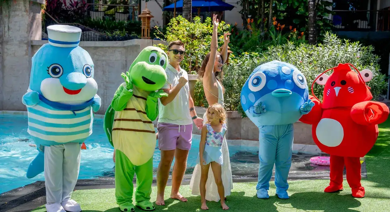 Familjefest i Thailand med barn och maskotar vid poolen. En glad familj njuter av sol och bad med en delfin, sk&ouml;ldpadda, fisk och krabba.