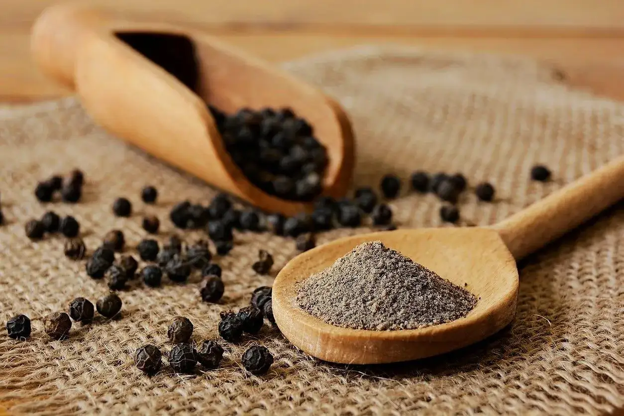 Cuchara de madera con pimienta negra molida, rodeada de granos enteros. Descubre las **pimienta negra propiedades** y su uso en la cocina.